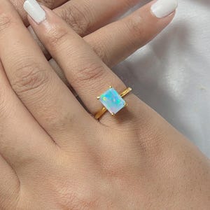 Könnte beinhalten: Ein goldener Ring mit einem rechteckigen Opal-Edelstein. Der Opal zeigt eine Mischung aus blauen, grünen und weißen Farben. Der Ring befindet sich an einer Hand mit weißem Nagellack.