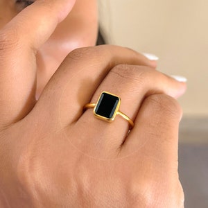Könnte beinhalten: Ein goldener Ring mit einem quadratischen schwarzen Edelstein. Der Ring befindet sich an einem Finger.