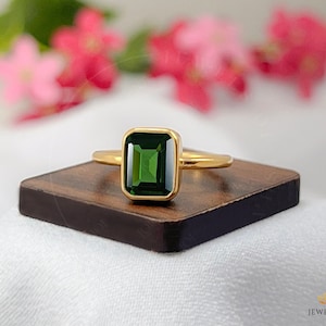 Puede incluir: Un anillo de oro con una piedra preciosa cuadrada de color verde esmeralda. El anillo está sobre una superficie de madera.