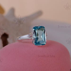 Pode incluir: Um anel de prata com uma grande pedra preciosa retangular azul clara. O anel está sobre uma superfície rosa, com um fundo desfocado. O design do anel inclui uma haste dividida e detalhes delicados ao redor da gema.