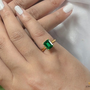 May include: A gold ring with a large, emerald-cut green gemstone. The ring has a simple gold band. The ring is shown on a hand with white nail polish.