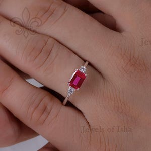 May include: A silver ring featuring a rectangular, deep red gemstone, flanked by two smaller, clear stones. The ring is set against a background of hands with turquoise nail polish. The ring is a piece of jewelry.