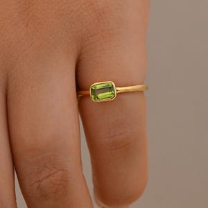 Può includere: Un anello in oro con una pietra preziosa di peridoto verde dal taglio rettangolare a smeraldo, fissata in una montatura a castone liscia. La fascia è sottile e lucida, con un design semplice che esalta il vibrante colore verde lime.