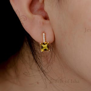 Puede incluir: Primer plano de un pendiente de oro con una gema amarilla cuadrada y una pequeña barra brillante. El pendiente está engastado en un marco dorado y se lleva en la oreja. El texto "Jewels of Isha" es visible.