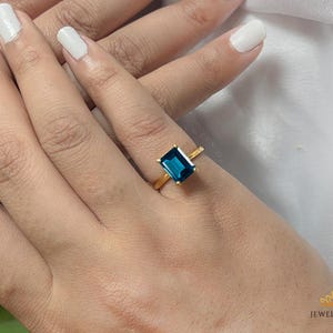 Könnte beinhalten: Ein goldener Ring mit einem rechteckigen, tiefblauen Edelstein. Der Ring befindet sich an einem Finger mit weißem Nagellack. Der Ring ist ein Schmuckstück.