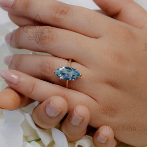 May include: A gold ring with a marquise-cut, light blue gemstone. The ring is set on a gold band and is displayed on a hand with manicured nails, surrounded by white flowers.
