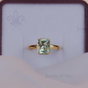 May include: A gold ring featuring a rectangular, light green gemstone set in a four-prong setting. The ring is displayed in a white jewelry box with a dark purple border, featuring the text Jewels of Isha printed in the corner and on the box.