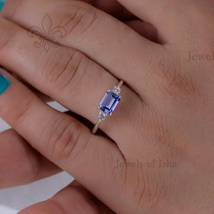 Puede incluir: Un anillo de plata con una piedra preciosa rectangular azul claro y piedras más pequeñas y claras. El anillo se muestra en una mano con esmalte de uñas turquesa. El anillo es de Jewels of Isha.