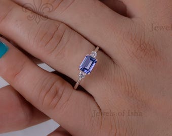 Puede incluir: Un anillo de plata con una piedra preciosa rectangular azul claro y piedras más pequeñas y claras. El anillo se muestra en una mano con esmalte de uñas turquesa. El anillo es de Jewels of Isha.