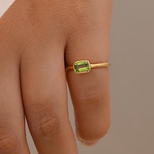 May include: A gold ring featuring a rectangular, emerald-cut green peridot gemstone set in a smooth bezel setting. The band is thin and polished, showcasing a simple design that highlights the vibrant lime green colour of the central stone.