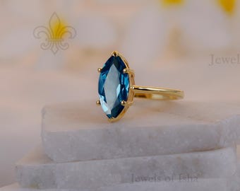 May include: A gold ring with a marquise-cut, blue gemstone. The ring is set on a white, layered surface, with a soft, blurred background. The ring's band is thin and gold, complementing the blue of the gemstone. The ring is a piece of jewelry.