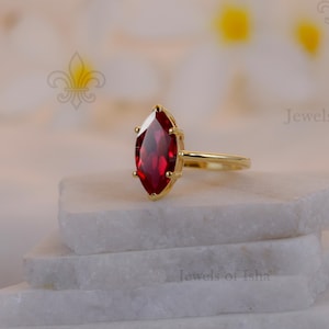May include: A gold ring with a marquise-cut, deep red gemstone. The ring is set on a white, layered surface, with a soft, blurred background. The ring's band is thin and delicate, and the gemstone is the focal point.