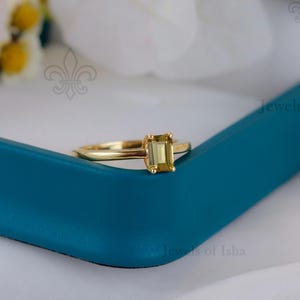 Peut inclure: Une bague en or avec une pierre précieuse rectangulaire vert clair. La bague est sertie dans un anneau en or et présentée dans un écrin bleu sarcelle. Le fond comprend des fleurs blanches.
