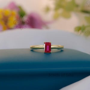 Peut inclure: Une bague en or avec une pierre précieuse rouge vif de forme rectangulaire. La bague est posée sur une surface bleu canard foncé, avec un tissu blanc doux et des fleurs floues en arrière-plan. C'est un bijou.