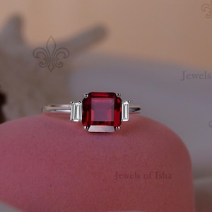 Pode incluir: Um anel de prata com uma grande pedra preciosa vermelha escura de corte quadrado como peça central, ladeada por duas pedras preciosas brancas retangulares menores. O anel é exibido em uma superfície rosa.