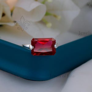 May include: A silver ring featuring a large, rectangular, deep red gemstone. The gemstone is the focal point, set against a simple silver band. The ring is displayed on a teal surface with white flowers in the background.