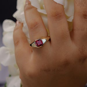 May include: A gold ring with a square, deep red gemstone set in a white bezel. The ring is on a person's finger, with white flowers in the background. The ring is a signet style.
