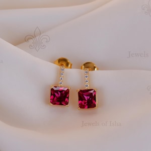 Peut inclure: Paire de boucles d'oreilles dorées avec des pierres précieuses carrées rouge rubis pendantes. Chaque boucle d'oreille a une petite barre sertie de pierres précieuses transparentes reliant la tige au carré rouge. Présentées sur un tissu crème drapé.