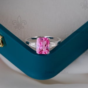 May include: A silver ring with a large, rectangular pink gemstone as the centerpiece. Two smaller, rectangular white gemstones flank the main stone. The ring is set against a teal blue jewelry box.
