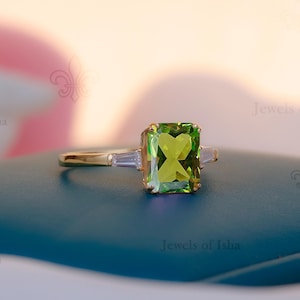May include: A gold ring featuring a large, rectangular, light green gemstone as the centerpiece, flanked by two smaller, rectangular, clear gemstones. The ring is set on a dark teal surface.