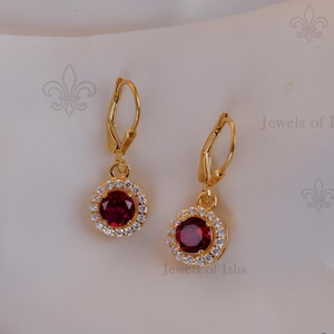 Può includere: Un paio di orecchini pendenti color oro. Ogni orecchino presenta una gemma rotonda di colore rosso intenso, circondata da gemme chiare più piccole. Gli orecchini sono sospesi da una chiusura a leva. Lo sfondo è un morbido tessuto bianco.