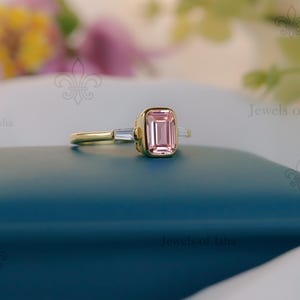 May include: A gold ring with a rectangular, pale pink gemstone as the main feature, with two smaller, rectangular, clear gemstones on either side. The ring is set on a blue surface, with blurred flowers in the background. The ring is a piece of jewellery.