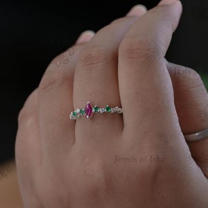 Peut inclure: Une bague en argent ornée d'une pierre rose centrale taille marquise, entourée de petites pierres rondes vertes et de pierres blanches transparentes sur l'anneau. La bague est portée au doigt et le texte Jewels of Isha est visible dans le coin.