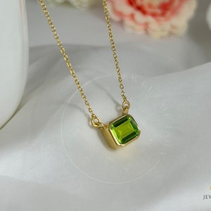 Può includere: Una collana color oro con un pendente rettangolare di pietra preziosa verde chiaro. Il pendente è incastonato in una lunetta dorata e appeso a una sottile catena d'oro. Lo sfondo è un morbido tessuto bianco.