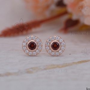 Könnte beinhalten: Ein Paar roségoldene Ohrstecker. Jeder Ohrring hat einen zentralen, runden, dunkelroten Edelstein, der von kleineren, klaren Edelsteinen umgeben ist. Der Hintergrund ist unscharf und zeigt den Text "Jewels of Isha".