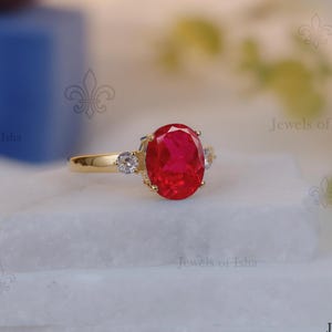 Puede incluir: Un anillo de oro con una gran gema roja ovalada como pieza central. Dos gemas claras más pequeñas flanquean la piedra principal. El anillo se exhibe sobre una superficie blanca.
