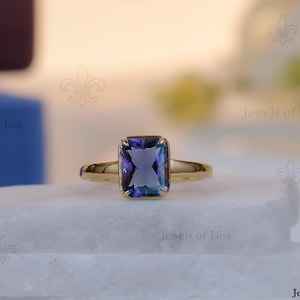 May include: A gold ring featuring a rectangular, multi-toned blue gemstone. The ring is displayed on a white surface, with the words "Jewels of Isha" visible in the background. The ring has a simple, polished gold band.
