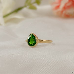 Puede incluir: Un anillo de oro con una piedra preciosa verde en forma de lágrima. El anillo está sobre un fondo blanco.