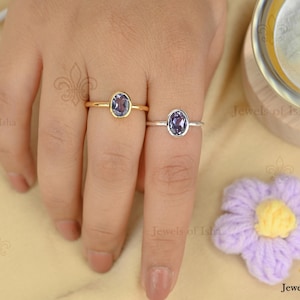 Könnte beinhalten: Zwei Ringe mit ovalen, lila Edelsteinen. Einer hat ein goldenes Band, der andere ein silbernes Band. Die Ringe werden auf einer Hand präsentiert, mit einer lila und gelben Blume im Hintergrund. Die Ringe sind von Jewels of Isha.