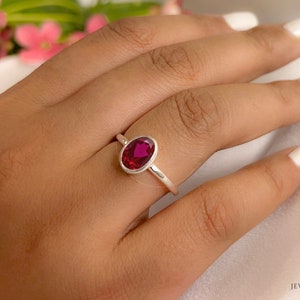 Könnte beinhalten: Ein silberner Ring mit einem leuchtend roten ovalen Edelstein. Der Ring befindet sich an einem Finger mit weißem Nagellack.