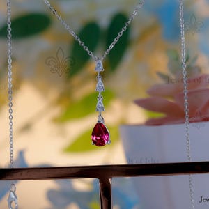 Peut inclure: Un collier en argent avec un pendentif en forme de goutte d'eau, orné d'une pierre précieuse rouge vif. Le pendentif est suspendu à une chaîne délicate, avec trois petites pierres précieuses claires au-dessus de la pierre principale. Le collier est exposé sur un présentoir.