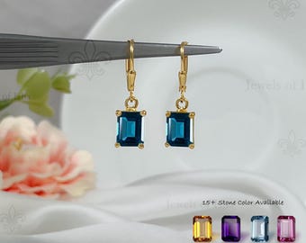 May include: A pair of gold-toned leverback earrings featuring rectangular, emerald-cut dark blue gemstones. The image includes text reading Jewels of Isha and 15+ Stone Color Available, with four additional small gemstone samples in yellow, purple, light blue, and pink.