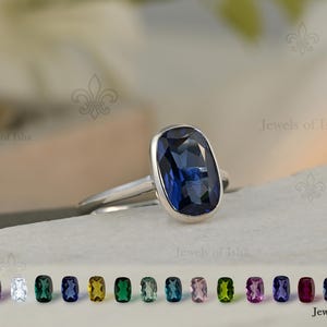 Pode incluir: Um anel de prata com uma grande pedra preciosa retangular azul escuro. O anel está sobre um fundo branco, com uma seleção de pedras preciosas coloridas abaixo. O anel é de Jewels of Isha.