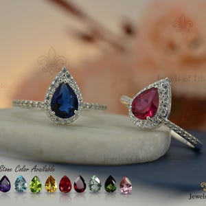 Puede incluir: Dos anillos de plata con piedras centrales en forma de pera, una azul profundo y otra rosa vibrante, rodeadas por un halo de pequeñas piedras claras. Los aros finos también están engastados con pequeñas piedras claras. Texto 15+ Stone Color Available.