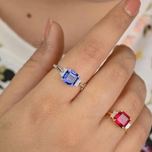 May include: A hand displays two rings. One ring features a blue emerald-cut gemstone with two baguette diamonds. The other ring has a red square gemstone with two baguette diamonds. Both rings are set on gold and silver bands.