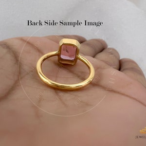 May include: A gold ring with a pink square gemstone. The ring is on a hand, and the back of the ring is visible.