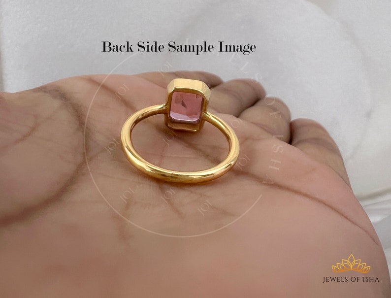 May include: A gold ring with a pink square gemstone. The ring is on a hand, and the back of the ring is visible.