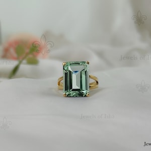 Puede incluir: Un anillo de oro con una gran piedra preciosa rectangular de color verde claro. El anillo tiene un diseño de caña dividida. El fondo es una tela blanca con un arreglo floral borroso.
