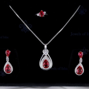May include: A jewellery set comprising a silver necklace with a teardrop-shaped red gemstone pendant, matching earrings, and a ring. The gemstones are surrounded by smaller, clear stones, set against a black background. The set is displayed in a black box.