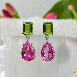 Op de afbeelding: Zilveren oorhangers met een rechthoekige, lichtgroene edelsteen aan de bovenkant en een peervormige, roze edelsteen aan de onderkant. De oorbellen worden getoond tegen een vage achtergrond met roze bloemaccenten.