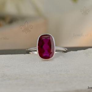 May include: A silver ring featuring a large, oval-shaped, deep pink gemstone set in a smooth bezel setting. The band is polished silver, and the ring is centered on a textured, light-colored surface. The text Jewels of Isha is visible.