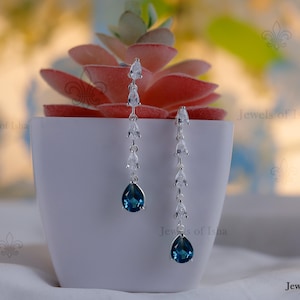 Peut inclure: Boucles d'oreilles pendantes en argent. Chaque boucle d'oreille présente une pierre précieuse bleu foncé en forme de goutte d'eau, avec de plus petites pierres transparentes le long de la chaîne. Les boucles d'oreilles sont présentées dans un pot blanc avec une plante succulente rouge. Le texte "Jewels of Isha" est visible.