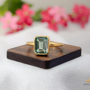 May include: A gold ring with a light green emerald-cut gemstone. The ring is on a wooden surface.