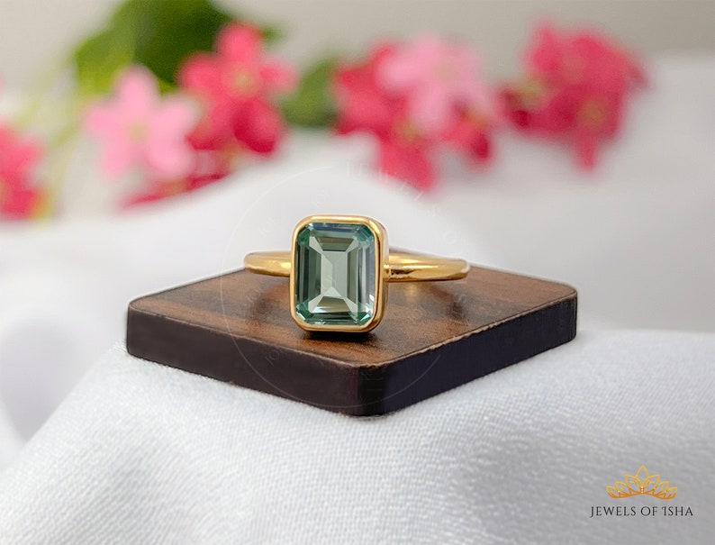 May include: A gold ring with a light green emerald-cut gemstone. The ring is on a wooden surface.