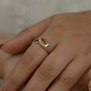 Peut inclure: Une bague en argent avec une pierre précieuse rectangulaire de couleur ambre. La bague est portée au doigt d'une personne, mettant en valeur le métal poli et la teinte chaude de la pierre. La bague est un bijou.