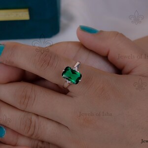 Op de afbeelding: Een zilveren ring met een grote, smaragdgeslepen groene edelsteen. Twee kleinere, heldere edelstenen flankeren de hoofdsteen. De ring zit aan een hand met turquoise nagellak, wat het ontwerp benadrukt.